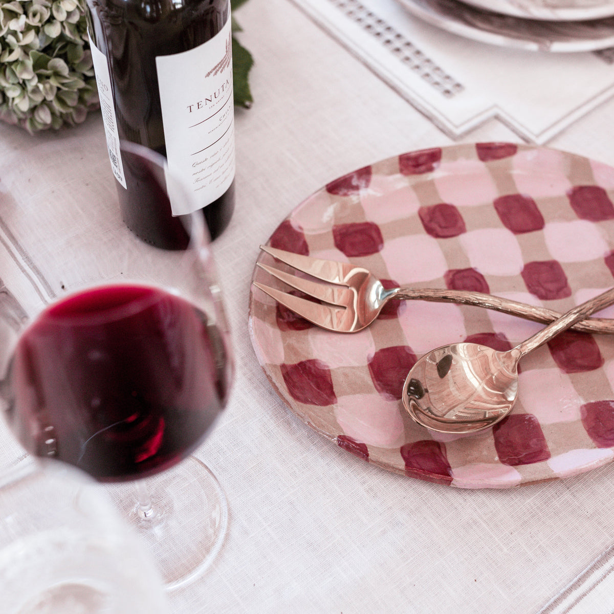 Wine bottle, glass, and checkered plate with cutlery on a table setting.